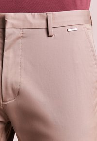 Pantaloni beige su misura con una texture liscia, caratterizzati da dettagli di cucitura sottili e un accento metallico del marchio sulla vita.