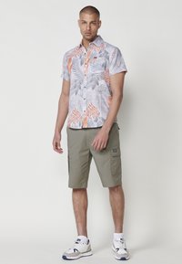 Camicia a maniche corte con bottoni, in tessuto con motivo di foglie tropicali grigie e arancioni, abbinata a pantaloni cargo olive e sneaker bianche.
