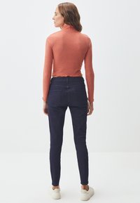 Rippiges, rostfarbenes Langarmoberteil mit einem Mock-Neck und geschwungenem Saum, kombiniert mit dunklen Skinny-Jeans und weißen Sneakers.