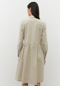Abito di cotone beige con maniche lunghe, chiusura a bottoni e vita arricciata, caratterizzato da una texture liscia e una silhouette fluida.