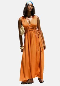 Robe maxi en tissu orange avec décolleté en V profond, taille froncée et accents floraux. Associée à un sac en osier et des sandales à lanières.