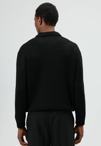 Zwarte gebreide trui met een brede geribde kraag en lange mouwen, met een ontspannen pasvorm en een gestructureerde stof, gedragen over donkere broek.