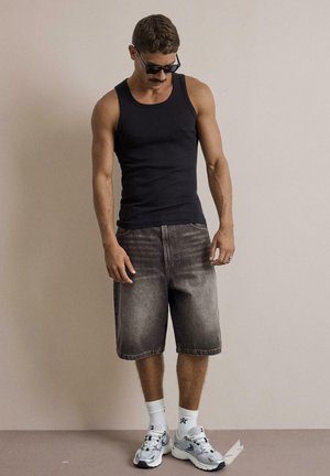 Mann trägt schwarzes Tanktop, ausgebleichte schwarze knielange Shorts, weiße Socken, silberne Sneakers und schwarze Sonnenbrille und steht vor einer schlichten Wand.