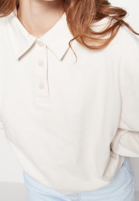 Polo de color beige claro con cuello, que presenta una tapeta de botones y una textura de tela suave, combinado con unos vaqueros de mezclilla azul claro.