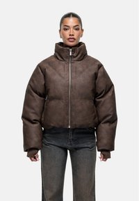 Vrouw met teruggetrokken haar, gekleed in een bruin gewatteerd jack met hoge kraag en lange mouwen, gecombineerd met donkere jeans.