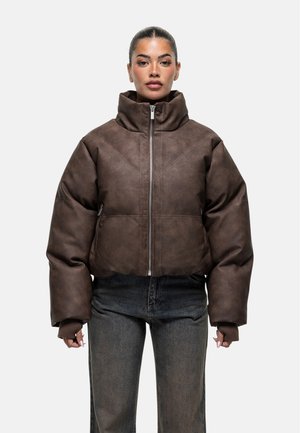 Vrouw met teruggetrokken haar, gekleed in een bruin gewatteerd jack met hoge kraag en lange mouwen, gecombineerd met donkere jeans.