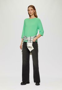 Grünes Langarmoberteil mit runder Halslinie, kombiniert mit weiten, dunklen Jeans. Ein kariertes Sweatshirt ist um die Taille gebunden und zeigt schwarz-weiße Muster.