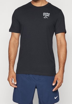 Homme portant un t-shirt Nike Trail noir et un short Nike bleu marine, debout avec les bras détendus le long du corps devant un fond uni.