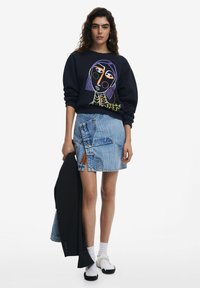 Zwarte sweatshirt met kleurraf grafiek, denim rok met asymmetrische snit, zwarte blazer, witte sokken en zwarte instappers.