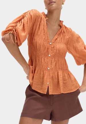 Femme portant un chemisier orange texturé avec manches bouffantes froncées à cordon et un short marron taille haute, posant les mains sur les hanches.
