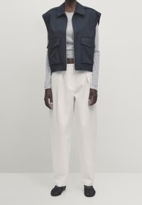 Granatowa krótka kamizelka z dwiema przednimi kieszeniami, w połączeniu z szarym, długim rękawem, białymi spodniami o szerokich nogawkach i czarnymi butami wsuwanymi. Gładkie tekstury.