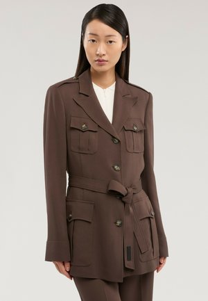 Junge Frau trägt ein braunes, mit Gürtel versehenes Blazer mit geknöpften Klapptaschen und einem weißen Top, steht vor einem einfarbigen hellen Hintergrund.