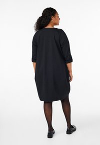 Schwarzes, knielanges Kleid mit dreiviertel langen Ärmeln und einem runden Ausschnitt. Es hat aufgerollte Ärmelsäume und eine Rückennaht-Detail. Kombiniert mit schwarzen Strumpfhosen und Schuhen.