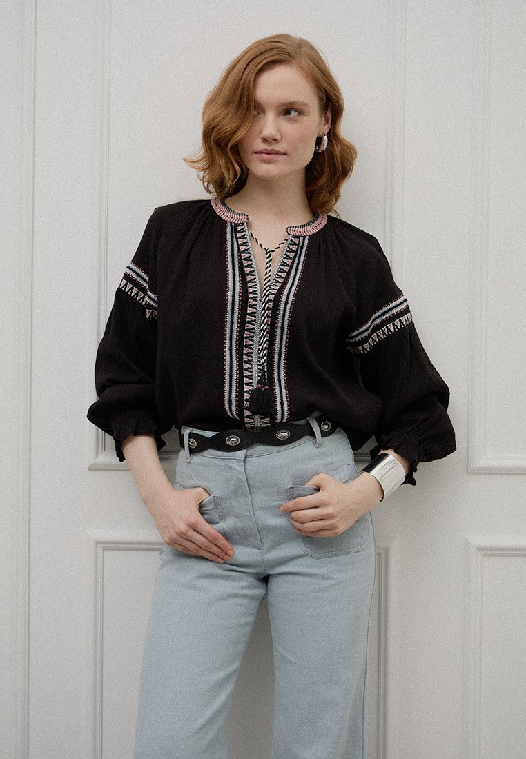 Schwarze Bluse mit bestickten Details, weit ausgestellten Ärmeln und V-Ausschnitt. Kombiniert mit hellblauen High-Waist-Hosen und einem schwarzen Gürtel.