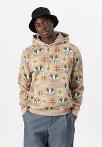 Giovane uomo che indossa un hoodie beige con motivi geometrici e floreali colorati, pantaloni blu e un cappello a secchiello nero su uno sfondo semplice.