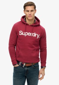 Burgundy hoodie with white "Superdry" logo, kangaroo pocket, and adjustable drawstring hood. Worn over plaid shirt and dark jeans.