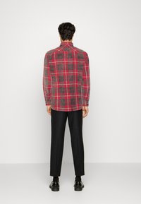 Camicia a quadri rossa e grigia con maniche lunghe, polsini con bottoni e colletto classico, realizzata in tessuto strutturato, indossata con pantaloni neri.