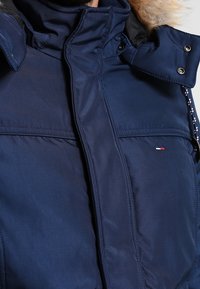 Mörkblå vinterjacka med hög krage, pälsfodrad huva, tryckknappar, dragkedjefodral och en liten röd, vit och blå logotyp på bröstet.