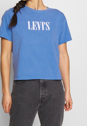 Nainen pukeutuneena siniseen Levi'sin lyhytpaitaiseen t-paitaan ja korkeavyötäröisiin mustiin farkkuihin, sivuletillä olkapään yli.