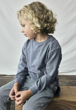 Barnet har på sig en grå, strukturerad sweatshirt med långa ärmar och rund halsringning, ihop med svarta och vita randiga byxor. Sitter på en träyta.