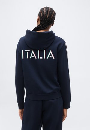 Person with braided hair wears navy blue hoodie and pants, "ITALIA" printed on back in white with green and red outlines.