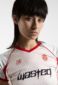 Balta sporta krekls ar sarkanajiem akcentiem, režģa rakstu un melnu logotipu "wasted." Satur sarkanu v-veida kakla izgriezumu un īsās piedurknes.