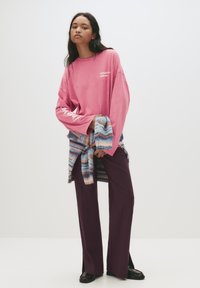 Tânără cu păr lung, închis la culoare, purtând un hanorac roz oversized, pantaloni purpurii, ținând un pulover tricotat cu dungi legat la talie.