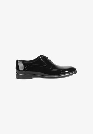 Chaussure habillée en cuir noir avec une finition laquée brillante, à lacets et à bout arrondi. Elle présente une semelle noire lisse et basse.