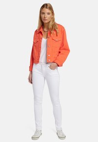 Giacca di denim arancione con due tasche sul petto e bottoni argentati, abbinata a un top bianco e jeans bianchi aderenti, completata da sneakers bianche.