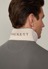 Sivi dukserica s mekom, strukturiranom tkaninom. Ima kontrastni svijetli ovratnik s "HACKETT" logotipom na stražnjoj strani. Jednostavan i moderan dizajn.