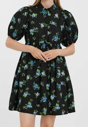 Femme portant une robe noire à imprimé floral bleu et blanc, manches courtes bouffantes, boutonnée devant et taille froncée avec ceinture à nouer.