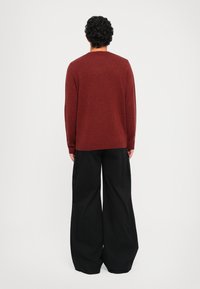 Roter Strickpullover mit rundem Kragen, kombiniert mit schwarzen weiten Cargo-Hosen, von hinten vor einem weißen Hintergrund gezeigt.