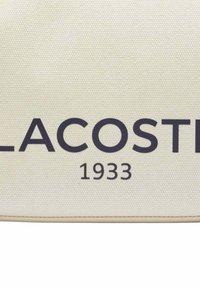 Sac en toile couleur crème avec un logo "LACOSTE" bleu foncé et "1933" en dessous, doté d'une surface texturée et sans matériel visible.