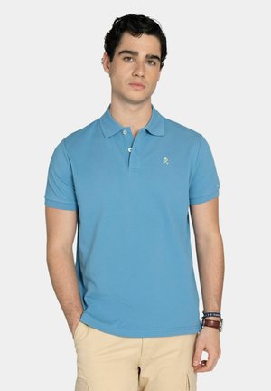 Hombre con camiseta polo de manga corta azul y pantalones cargo beige, de pie con una mano en el bolsillo contra un fondo liso.