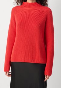 Rode gebreide trui met een hoge hals, geribbelde textuur en lange mouwen. Heeft een ontspannen pasvorm en een effen kleur zonder patronen.