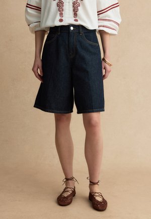 Vrouw draagt donkerblauwe denim shorts tot op de knie, wit blouse met rode borduursels en bruine veterplatte schoenen, staand tegen een beige achtergrond.