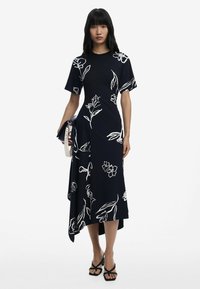 Dunkelblaues Kleid mit weißen Blumenmustern, kurzen Ärmeln und asymmetrischem Saum. Kombiniert mit schwarzen High Heels Sandalen und einer Clutch.