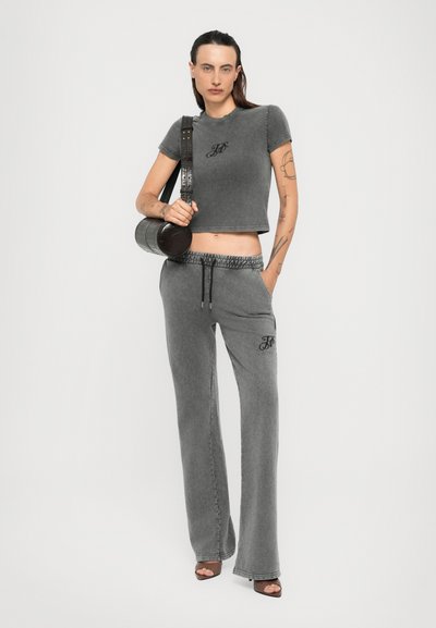 Jong persoon met tatoeages, gekleed in een grijze cropped t-shirt, bijpassende wijde broek, bruine haksandalen en met een zwarte cilindervormige schoudertas.
