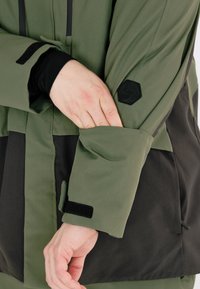 Grüne und schwarze Jacke mit langen Ärmeln, strukturierter Stoff, verstellbare Manschetten mit Klettverschluss und ein sechseckiges Logo am Ärmel.