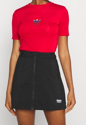 Personne portant un haut rouge à manches courtes avec un logo Adidas arc-en-ciel et une jupe noire avec une fermeture éclair devant et une étiquette Adidas.