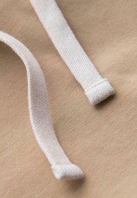 Detailní záběr na dvě bílé tkané šňůrky kapuce na béžovém pozadí.