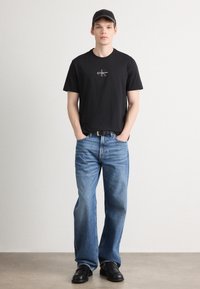 T-shirt preta de algodão com logótipo, jeans de ganga azul, cinto preto e sapatos pretos. O modelo está de mãos nos bolsos contra um fundo neutro.