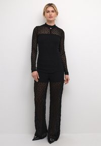 Schwarzer Spitzen-Bodysuit mit langen Ärmeln und hohem Kragen. Durchsichtige, gemusterte Textur mit einem Schlüsselloch-Detail am Ausschnitt. Eng anliegende Shorts darunter.