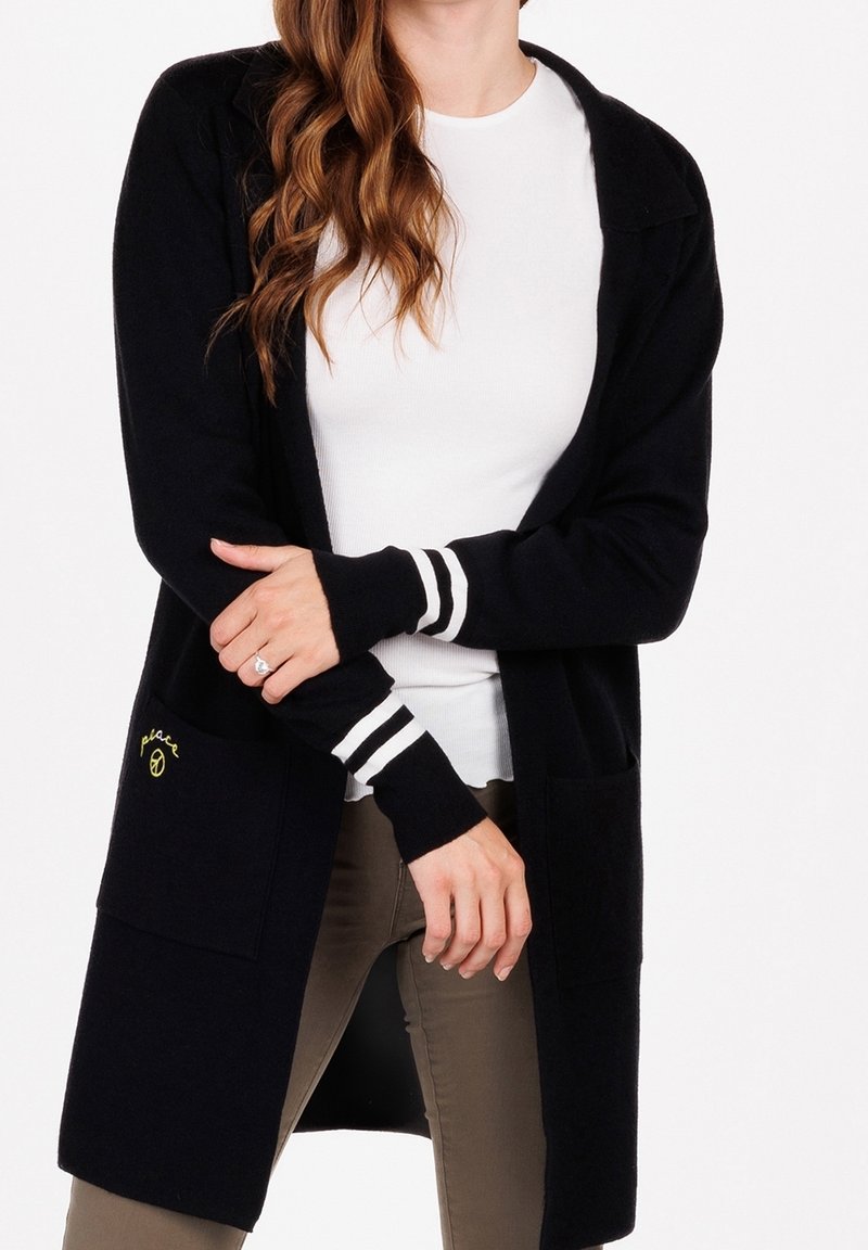 Czarny kardigan z długimi rękawami, białe paski na mankietach, dwie przednie kieszenie, miękka tekstura oraz naszywka z logo na lewej kieszeni.