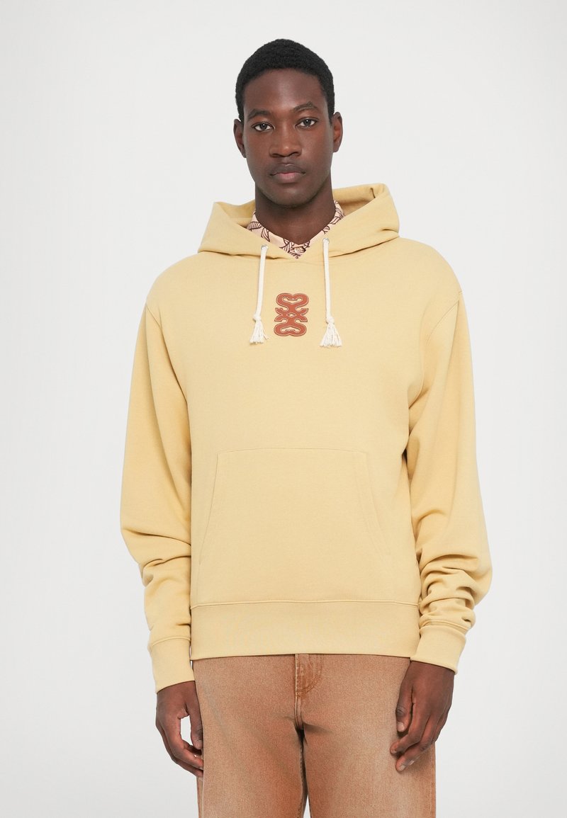 Mann iført en beige hettegenser med hvite snorer og et sentralt rødt abstrakt logo, sammen med lysebrune jeans mot en ensfarget bakgrunn.