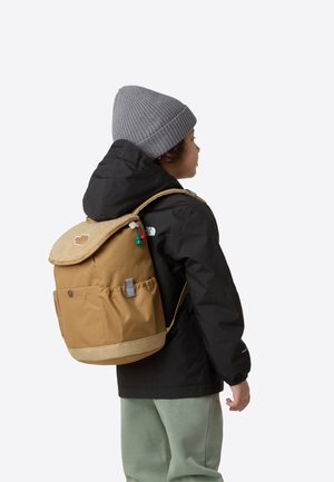 Sac à dos marron avec un rabat texturé beige, une poche latérale et des accents verts. Porté par un enfant en veste noire et bonnet gris.