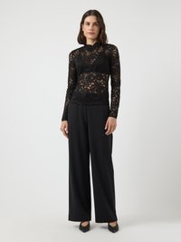 Top lungo a maniche nere in pizzo con scollatura alta, motivo floreale e sezioni trasparenti, abbinato a pantaloni neri a gamba larga e mocassini con punta.