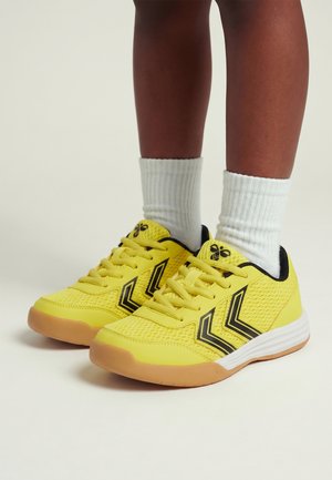 Gambe con calzini bianchi a coste e scarpe da ginnastica giallo brillante con strisce a chevron nere e suole in gomma su sfondo chiaro.