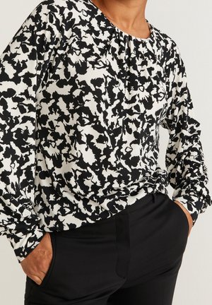 Person iført en sort og hvid mønstret bluse med lange ærmer, stoppet ned i sorte bukser, med hænderne i lommerne mod en ensfarvet baggrund.