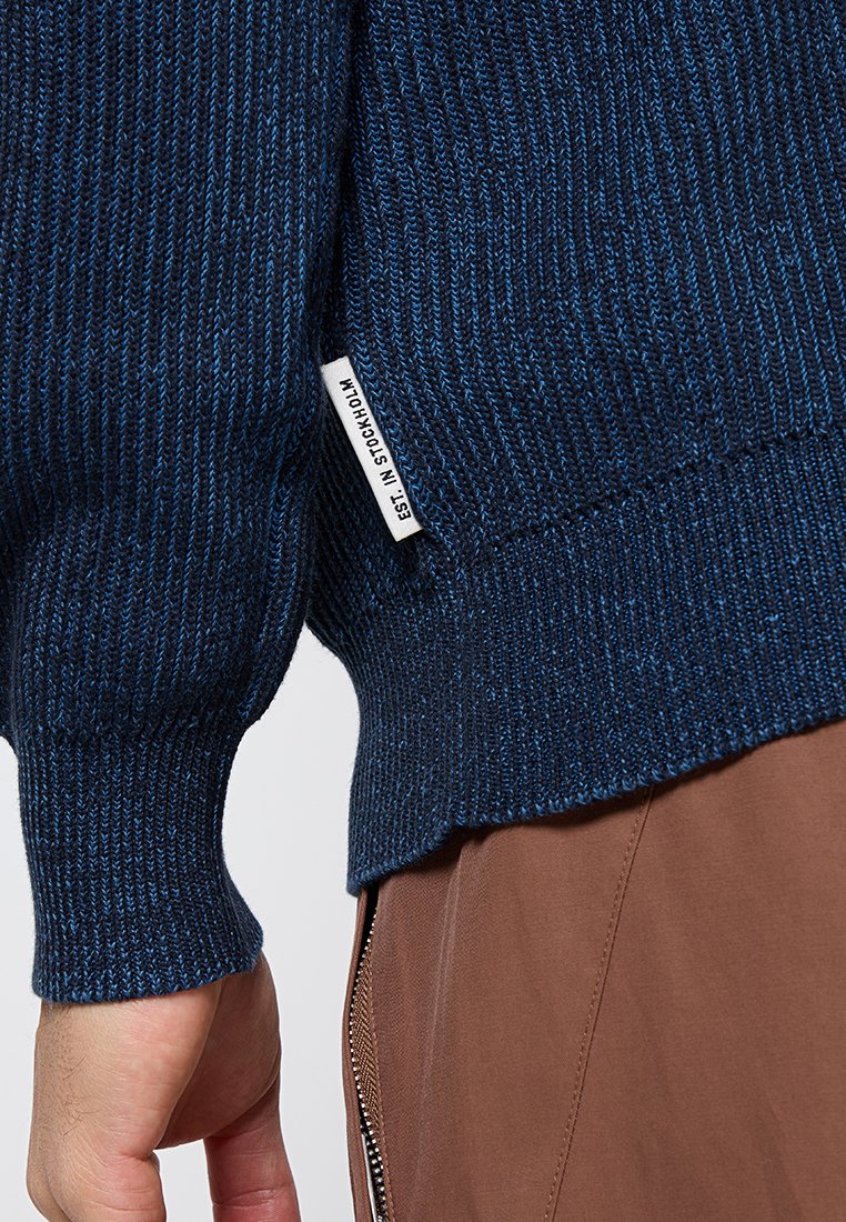 Nahaufnahme einer Hand in einem blauen Strickpullover mit einem weißen Etikett, auf dem "EST. IN STOCKHOLM" steht, kombiniert mit braunen Hosen, die einen sichtbaren Reißverschluss aufweisen.